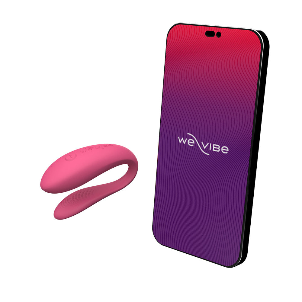 c-shaped wearable sex toy in pink next to a cell phone with a we-vibe logo on its screen. meant to show that the sex toy can be controlled via the phone app