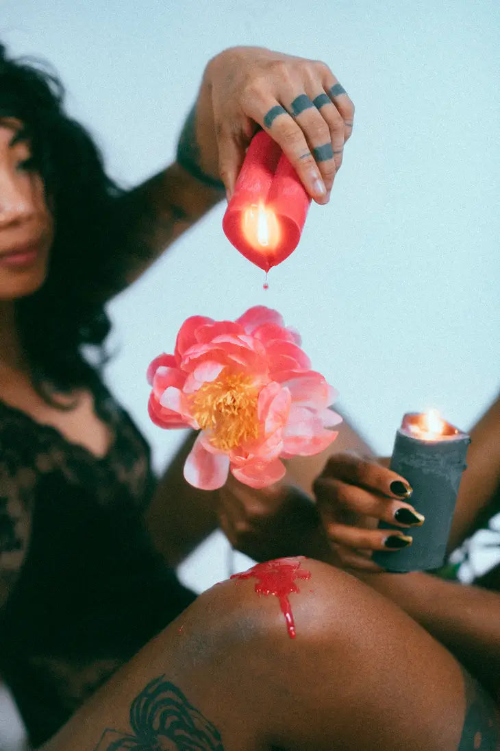 in foreground a lit red pillar candle is dripped onto brown person's knee. another hand holds a lit black candle. 